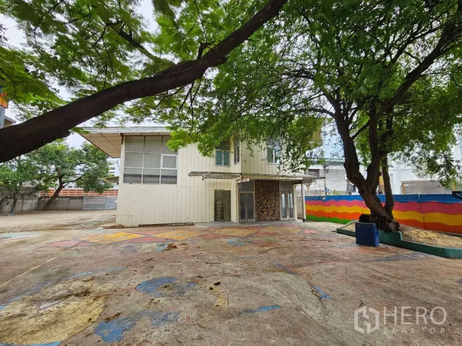 独栋房屋 for rent in Watthana, Bangkok - 两层独栋住宅外观，配有宽阔铺装庭院、成荫大树和彩色围墙。