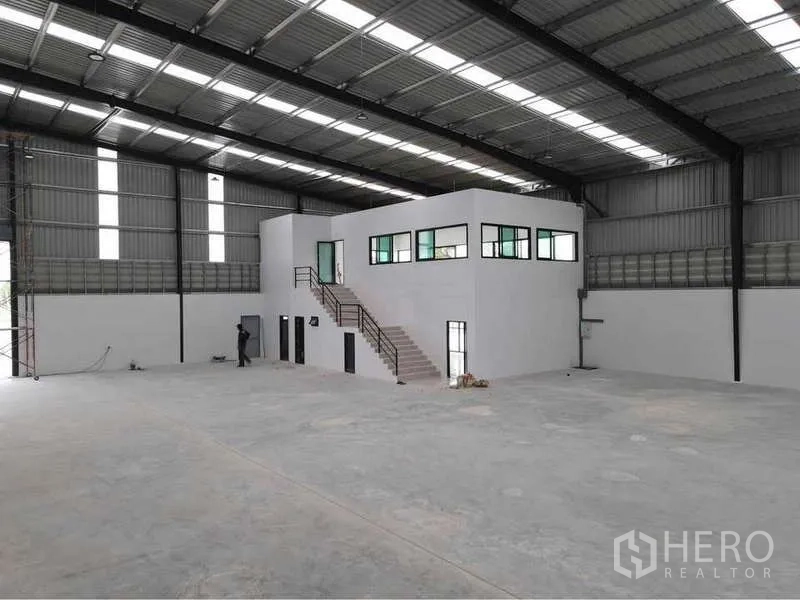 Warehouse for rent or sale in Nikhom Phatthana, Rayong - Spacious warehouse interior with high steel roof, polished concrete floor, and a white two-storey office block with windows and stairs.