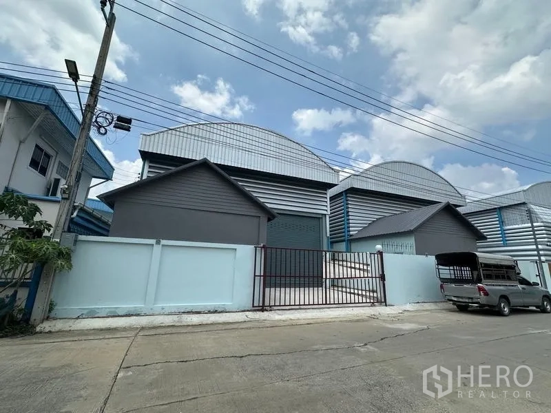 Factory for rent in Pak Kret, Nonthaburi - Street view of a modern mini factory with arched metal roofs, light blue perimeter wall and gated entrance in an industrial lane.