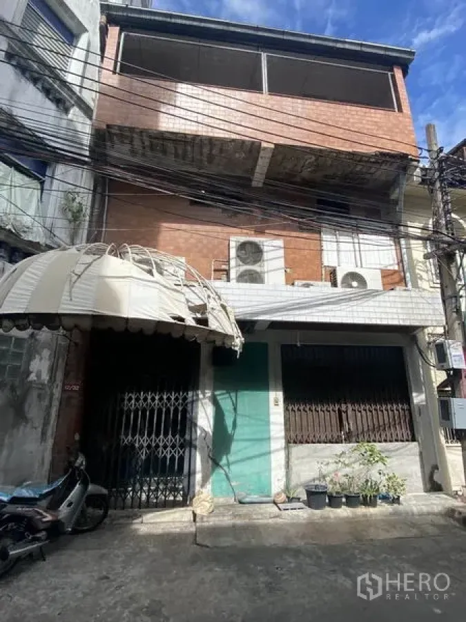 Shophouse for sale in Khlong Toei, Bangkok - Rear lane view of the shophouse with metal gates, canopy and large external AC units under a blue sky in Khlong Toei, Bangkok.