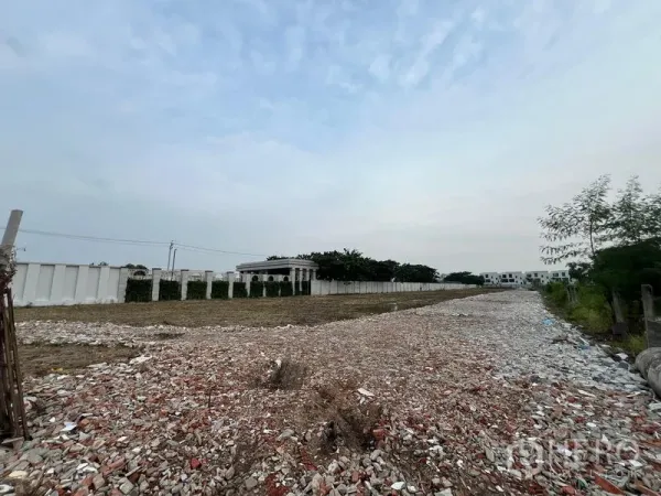 地 出租在Lat Krabang, Bangkok - 叻甲挽长条形地块，地面铺碎石，周边有围墙与绿树，天空多云。