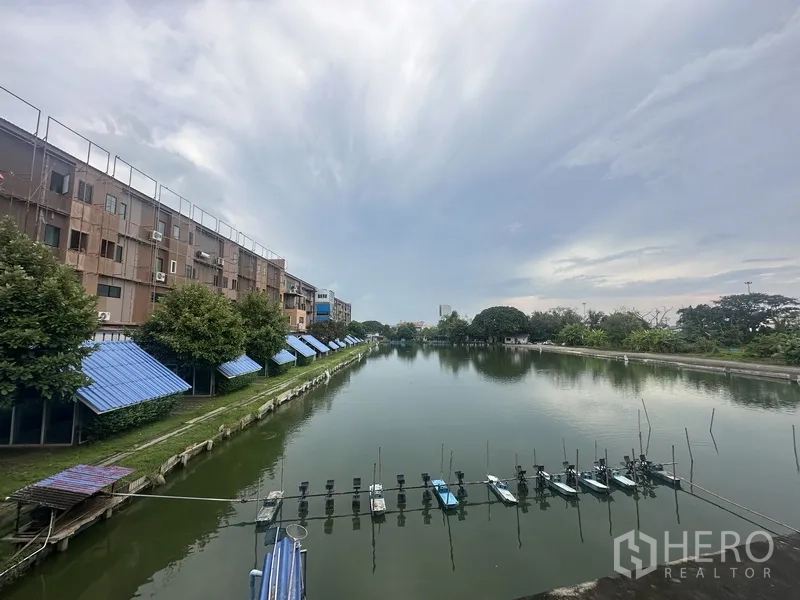 Standalone for rent in Prawet, Bangkok - View of a large fish pond bordered by buildings and trees under a cloudy sky.