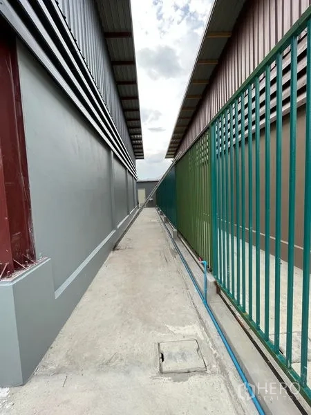 Warehouse for rent in Khlong Sam Wa, Bangkok - Narrow service lane between neighboring warehouses with green fence, water pipe, and drainage covers.
