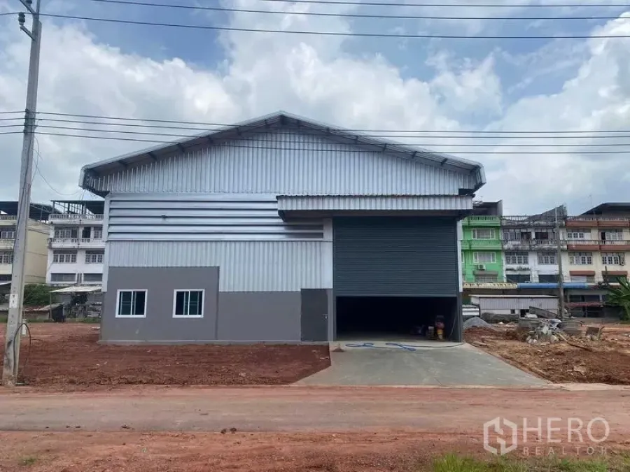 Warehouse for rent in Bang Bon, Bangkok - Front view of the warehouse with large roller shutter, driveway ramp and nearby shophouses.