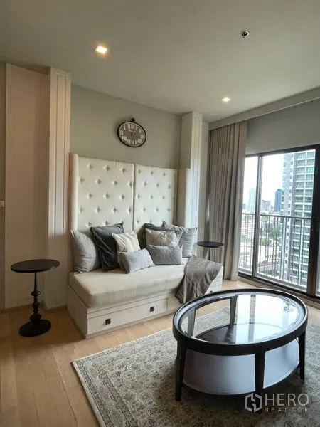 Condominium for rent in Bangkok - Bright living area with tufted wall sofa, round coffee table, wood floors, and a balcony city view in a Khlong Toei condo.