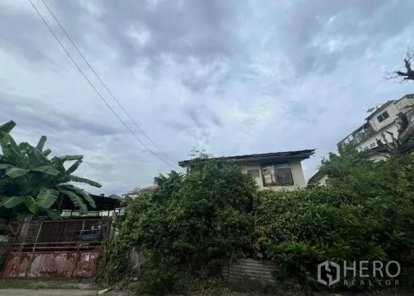 Land for sale in Phra Khanong, Bangkok - Overgrown land plot with an old two-storey house and a metal gate under a cloudy sky.
