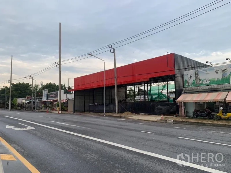 Warehouse for rent in Bueng Kum, Bangkok - Angled view of a roadside warehouse with red cladding, multi-lane road, streetlights and nearby shops.