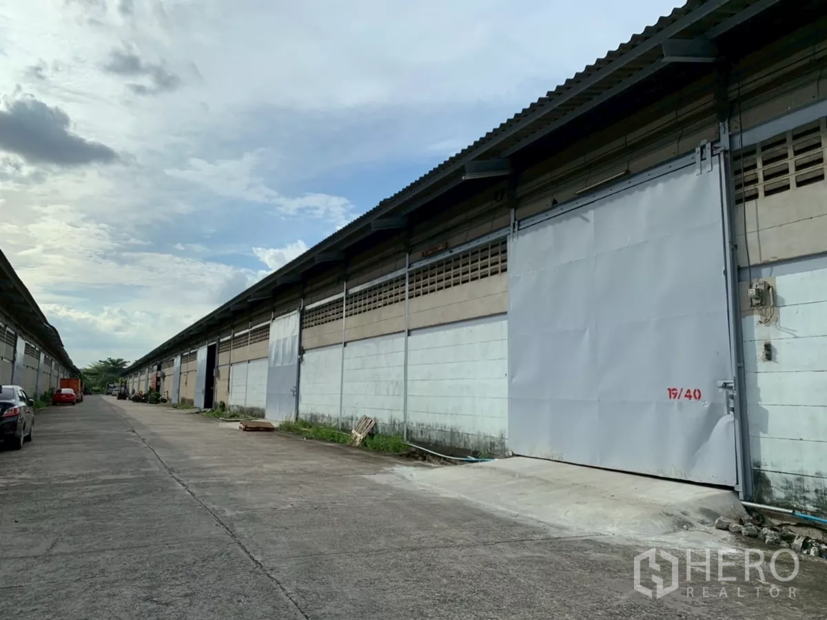 Warehouse for rent in Bang Phli, Samut Prakan - Row of industrial warehouses with large metal sliding doors and a wide driveway for truck access.