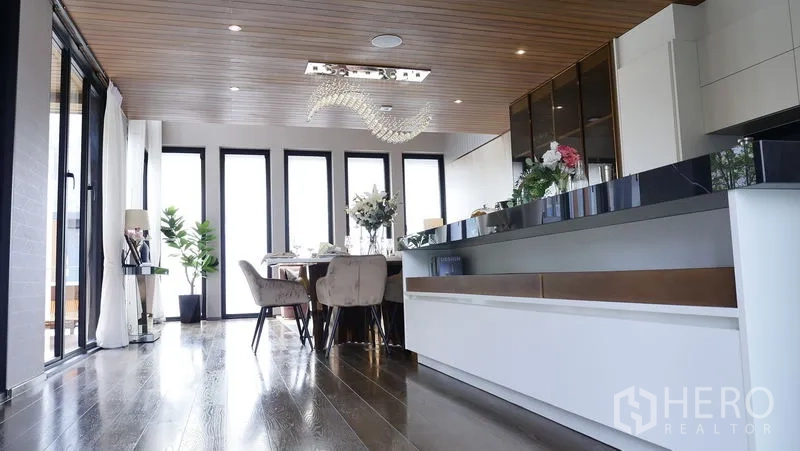 Detached House for sale in Prawet, Bangkok - Open‑plan kitchen and dining with long island, wood ceiling and tall windows.