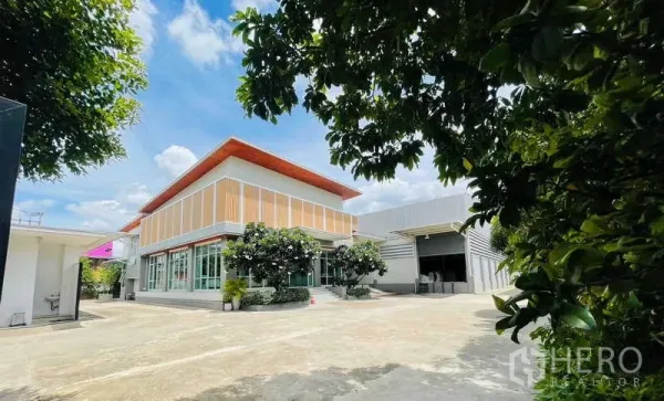 Factory for sale in Krathum Baen, Samut Sakhon - Modern two-storey office and showroom beside a metal factory with a wide concrete yard and trees.
