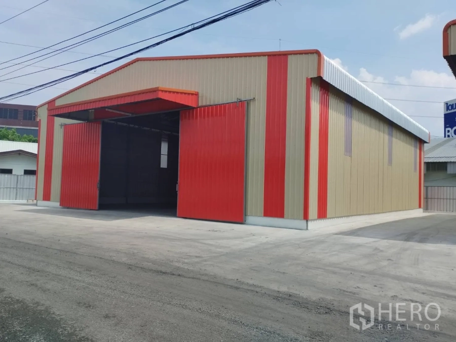 Warehouse for rent in Bang Khen, Bangkok - Angled exterior of the warehouse showing red sliding door, beige metal siding, and street access.