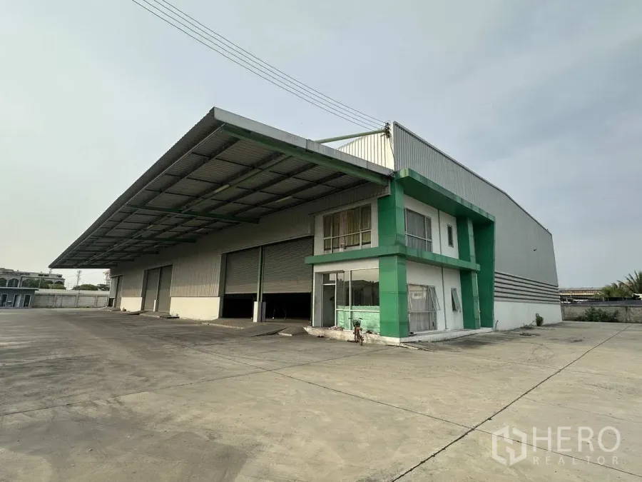 Factory for rent in Mueang Samut Prakan, Samut Prakan - Standalone factory exterior with green-trimmed office block, wide loading canopy, multiple roller shutters and a large concrete yard.