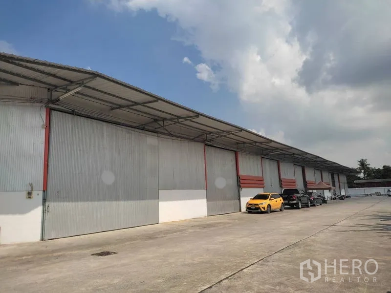Warehouse for rent in Pathum Thani - Long warehouse block with vehicles parked along a broad concrete forecourt.
