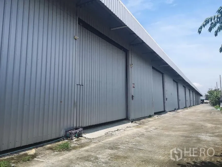 Warehouse for rent in Lam Luk Ka, Pathum Thani - Exterior row of metal-clad warehouse bays featuring large sliding doors and a driveway for truck access.