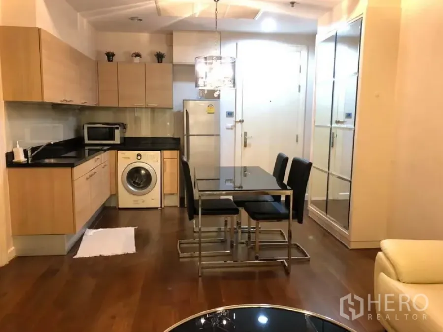 Condominium for rent in Pathum Wan, Bangkok - Open-plan kitchen with light wood cabinets, black countertops, appliances, washing machine and a glass dining table under a chandelier.