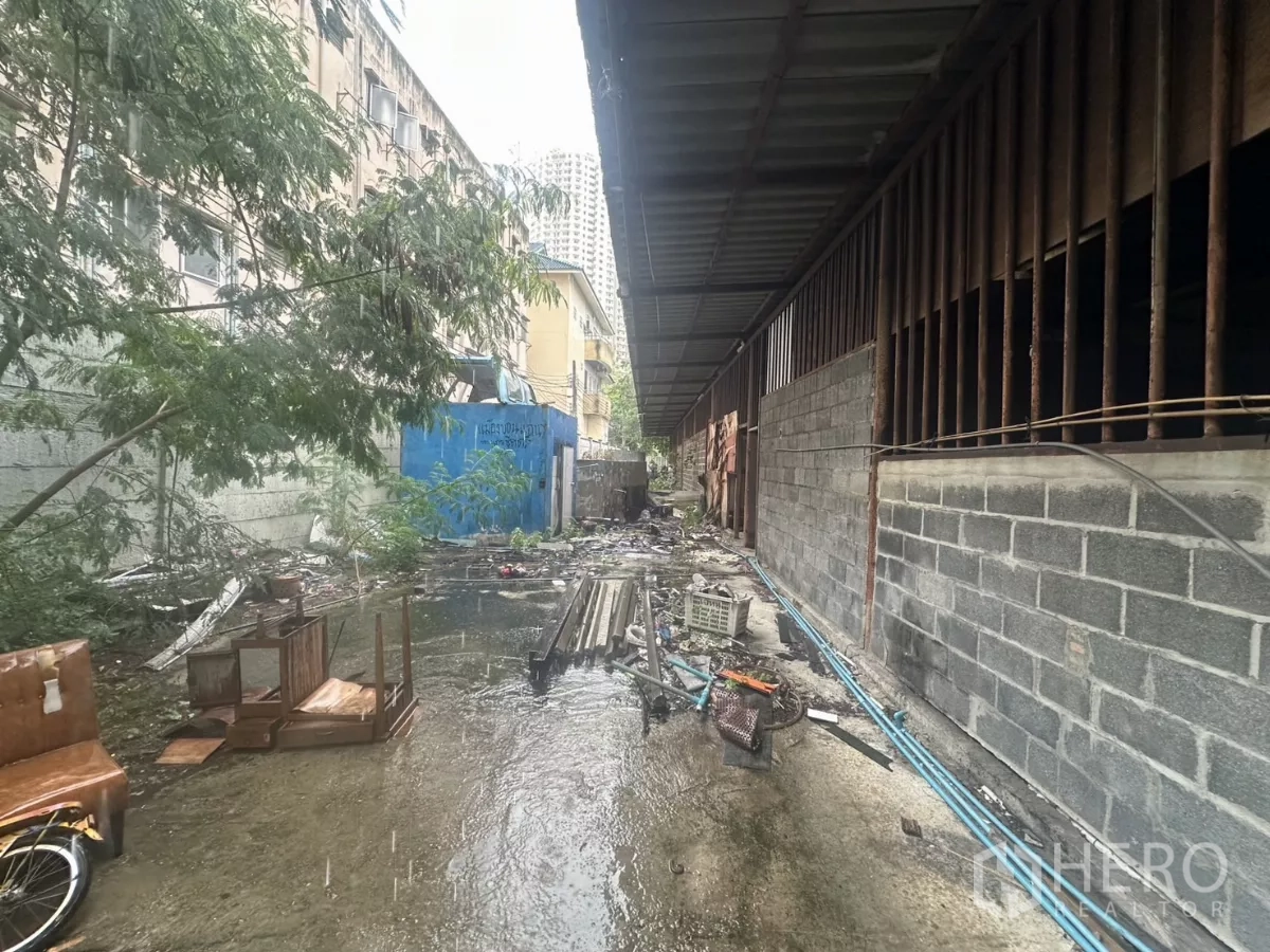 Warehouse for rent or sale in Yan Nawa, Bangkok - Rear alley beside the warehouse showing drainage water, trees and materials stacked along the wall.