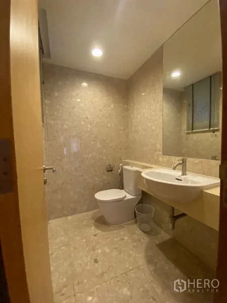 Condominium for rent in Watthana, Bangkok - Compact guest bathroom with wall-mounted sink, toilet, and large mirror over stone tiles.