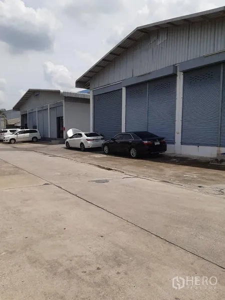 货栈 出租在Bang Phli, Samut Prakan - 一排带卷闸门的仓库建筑沿宽混凝土道路分布。