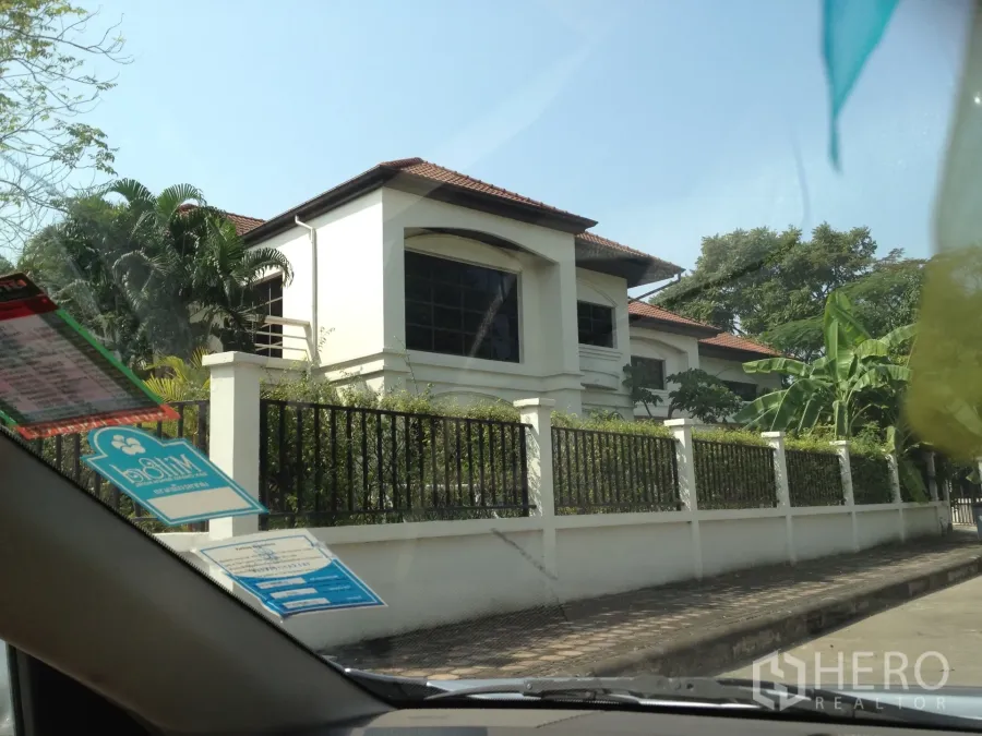 Detached House for rent in Nonthaburi - Two-storey white detached house with terracotta tile roof, large windows and a black metal fence amid lush trees in Pak Kret, Nonthaburi.