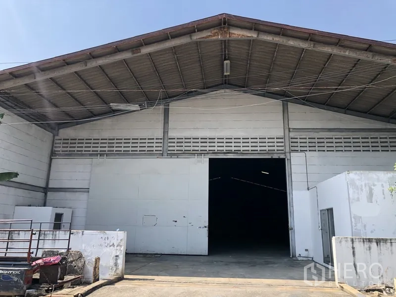 Warehouse for rent in Khlong Luang, Pathum Thani - Front exterior of a high-gable warehouse with metal roof, wide sliding door and concrete loading apron in an industrial area.