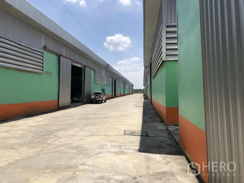 Warehouse for rent in Pathum Thani - Wide concrete driveway between green-and-orange warehouse blocks with metal cladding and truck access.