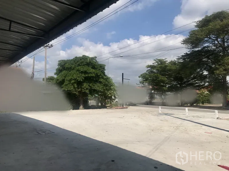 Warehouse for rent in Lam Luk Ka, Pathum Thani - Large paved forecourt and street access with trees and power lines in front of the warehouse.