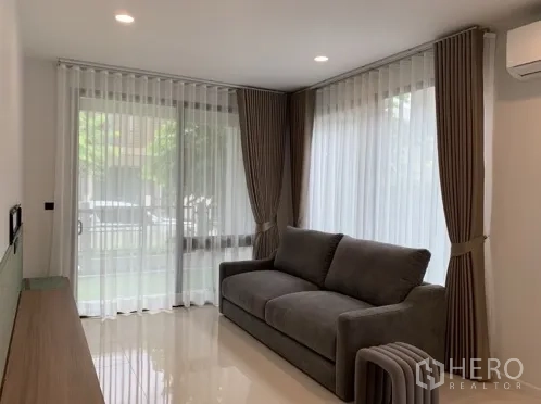 Detached House for rent in Bang Kapi, Bangkok - Cozy living room with grey sofa, corner windows and floor-to-ceiling drapes.