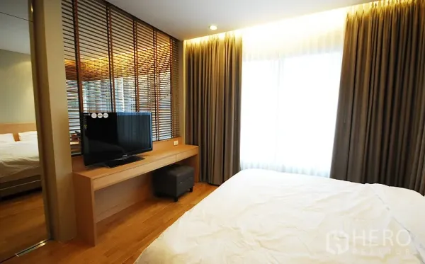 Condominium for rent in Khlong Toei, Bangkok - Bedroom with TV on wooden console, slatted partition, floor-to-ceiling curtains and warm ambient lighting.