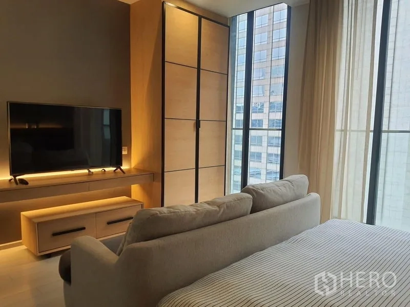 Condominium for rent in Pathum Wan, Bangkok - Corner bedroom with wardrobe, TV and floor-to-ceiling windows showcasing a city view.