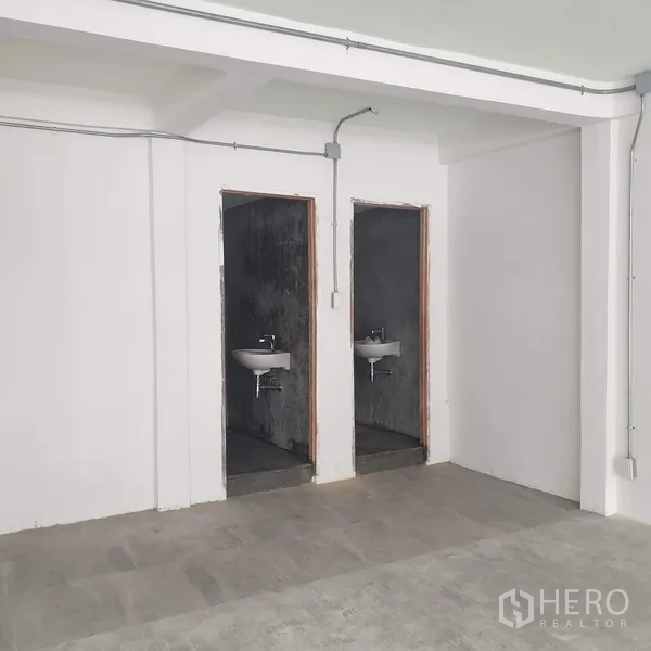 Home Office for rent in Khlong Toei, Bangkok - Two minimalist restrooms with white wall-mounted sinks and raw concrete walls.