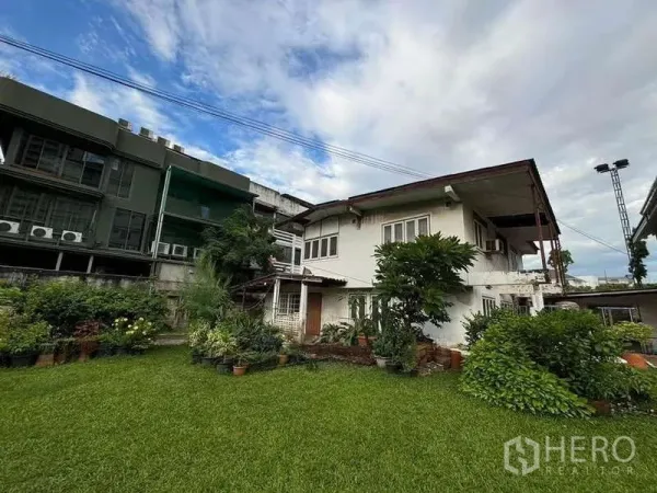 独立式住宅 出租在Suan Luang, Bangkok - 两层独栋房屋，宽阔草坪与成树，摆放花盆，城市环境下的多云天空。