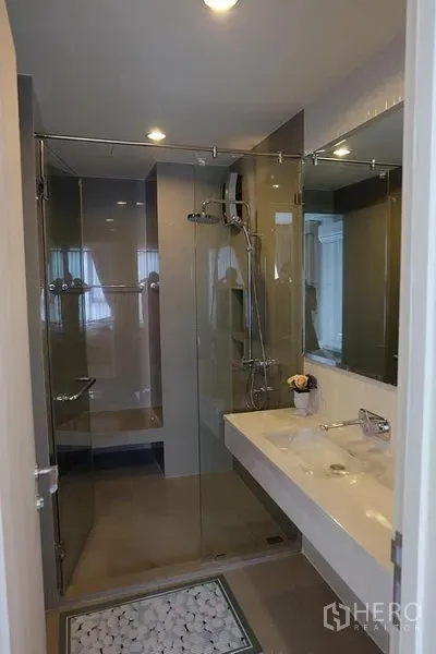 Condominium for rent in Khlong Toei, Bangkok - Modern bathroom featuring a glass-enclosed shower and long white vanity with mirror.