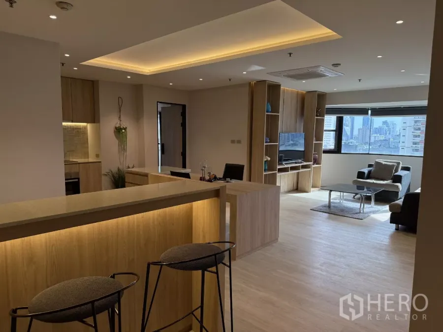 Condominium for rent in Watthana, Bangkok - Open-plan living area with wood breakfast bar, built-in TV wall, sofa seating and a wide window with city views.