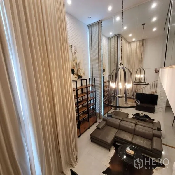 Condominium for rent in Khlong Toei, Bangkok - Double-height living room with floor-to-ceiling curtains, leather sectional, and modern chandeliers.