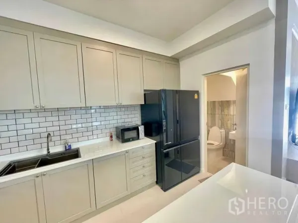Townhouse for rent or sale in Prawet, Bangkok - Modern L‑shaped kitchen with subway tile backsplash, tall cabinets and a black fridge beside the guest bathroom.