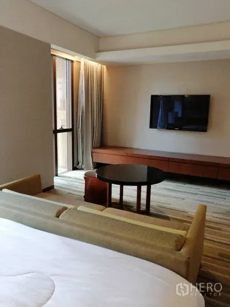 Condominium for rent in Pathum Wan, Bangkok - Cozy living area with wall-mounted TV, balcony door and curtains beside the bed.