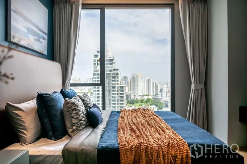 Condominium for rent in Khlong Toei, Bangkok - Bedroom city view framed by large window with layered pillows and throw