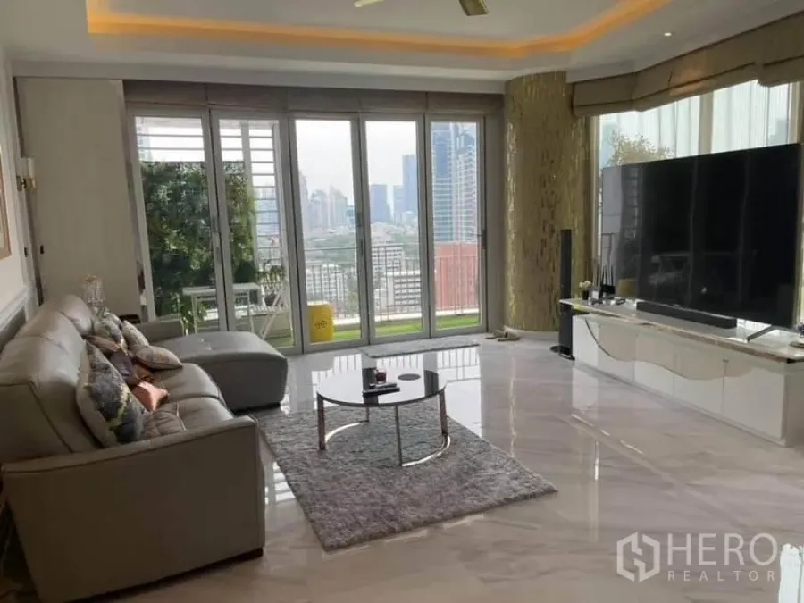 Condominium for sale in Pathum Wan, Bangkok - Bright living room with marble floors, L‑shaped sofa, and floor‑to‑ceiling windows opening to a city‑view balcony.