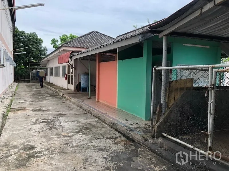 Factory for sale in Lam Luk Ka, Pathum Thani - Service walkway alongside single-storey auxiliary buildings with pastel walls and canopy.