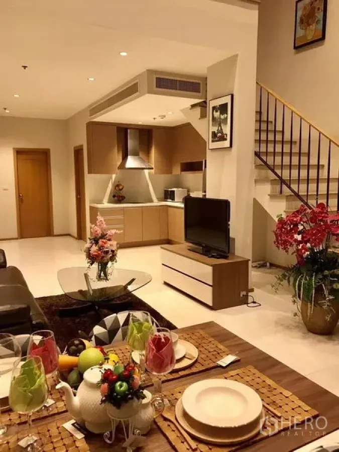 Condominium for rent in Khlong Toei, Bangkok - Dining setup facing living room with TV, open kitchen and staircase in a warm-toned condominium.
