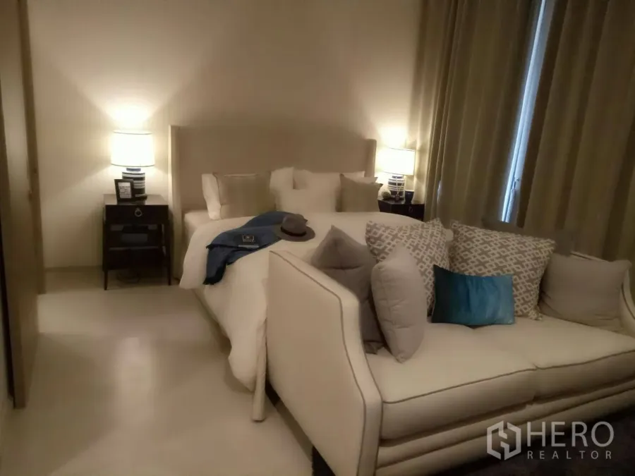 Condominium for rent or sale in Pathum Wan, Bangkok - Cozy bedroom with a plush sofa, bedside tables with lamps, and floor-length curtains in neutral tones.