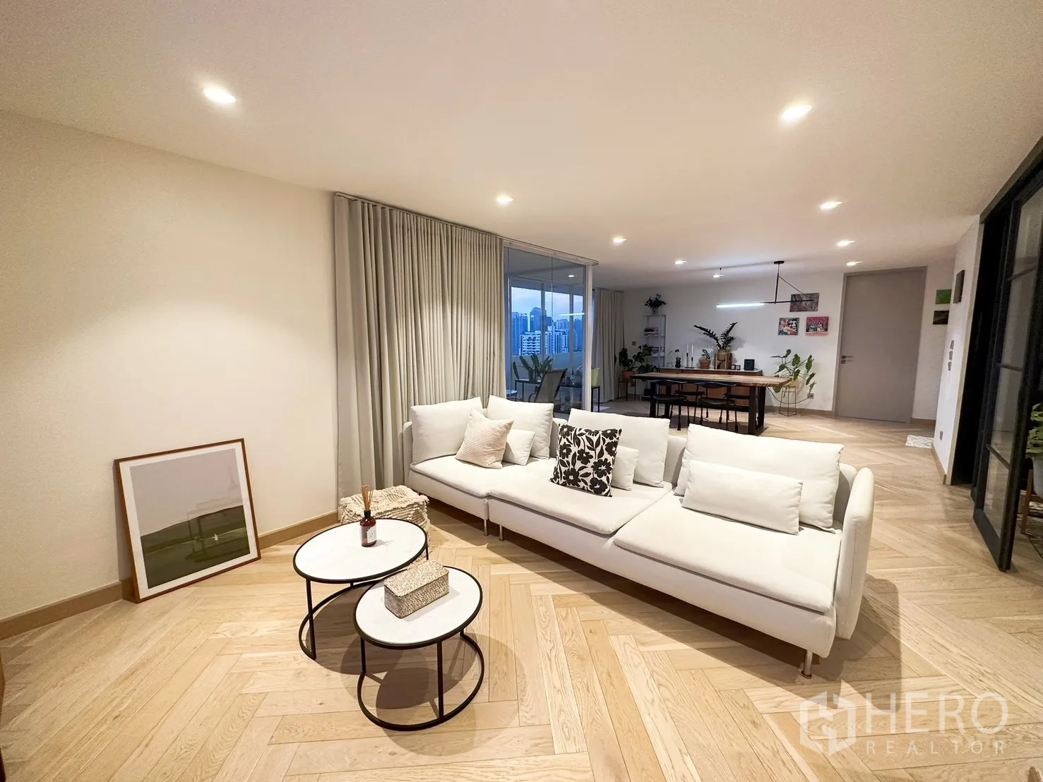 Condominium for rent or sale in Watthana, Bangkok - Open-plan living room with white sofa, herringbone wood floors, and glass walls to the city-view dining area.