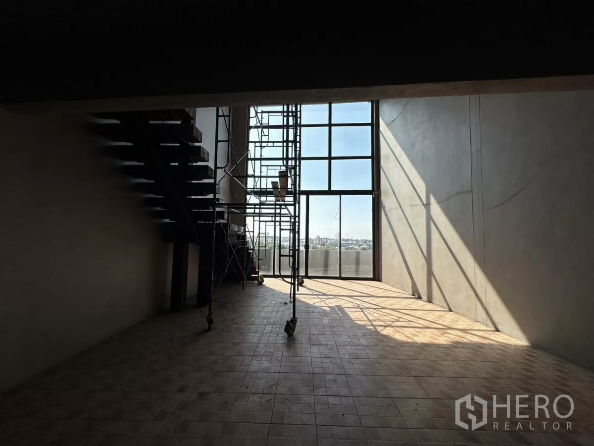 Shophouse for rent in Bang Na, Bangkok - Double‑height glass wall with scaffolding casting shadows across the tiled floor.