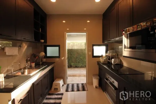 Detached House for rent in Prawet, Bangkok - Sleek galley kitchen with dark cabinets, built-in appliances and a door opening to the garden.
