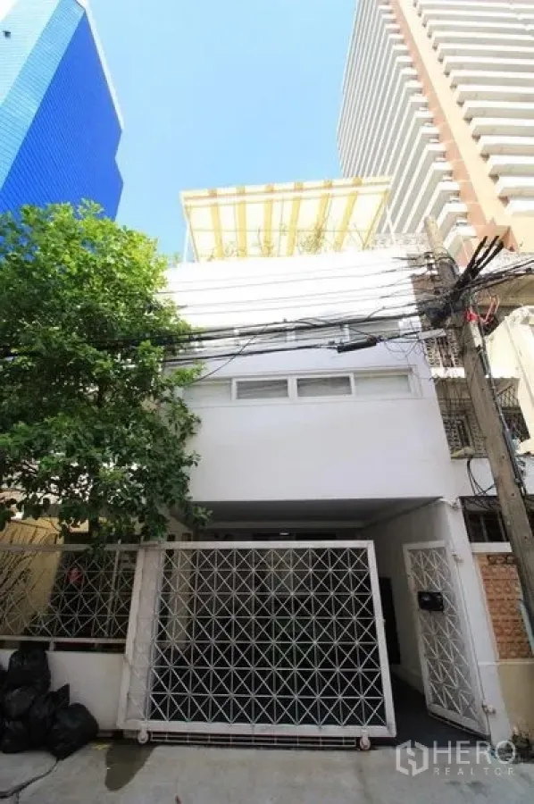 Townhouse for rent in Watthana, Bangkok - White townhouse facade with lattice gate, roadside tree and surrounding high-rises.