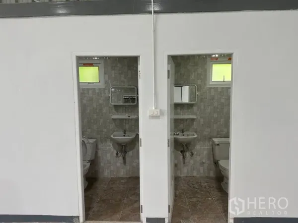 Warehouse for rent in Khlong Toei, Bangkok - Two tiled restrooms with toilets, sinks, mirrors, and small windows.