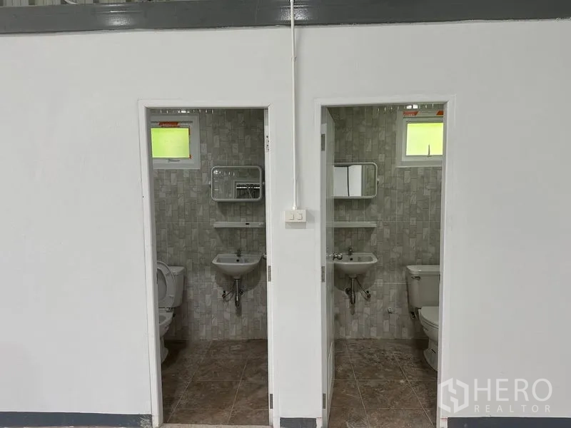 Warehouse for rent in Khlong Toei, Bangkok - Two tiled restrooms with toilets, sinks, mirrors, and small windows.