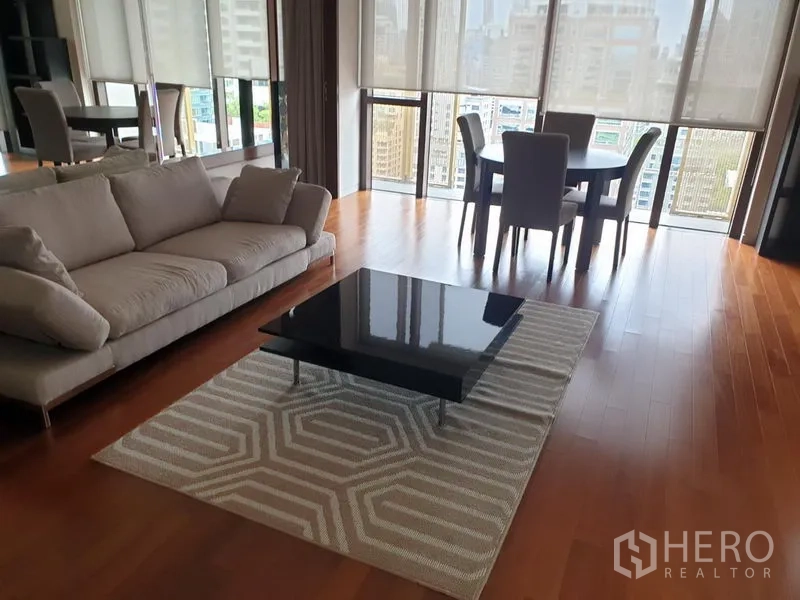 Condominium for rent in Pathum Wan, Bangkok - Bright lounge with sofa, black coffee table on rug, dining set and panoramic windows overlooking Bangkok skyline.