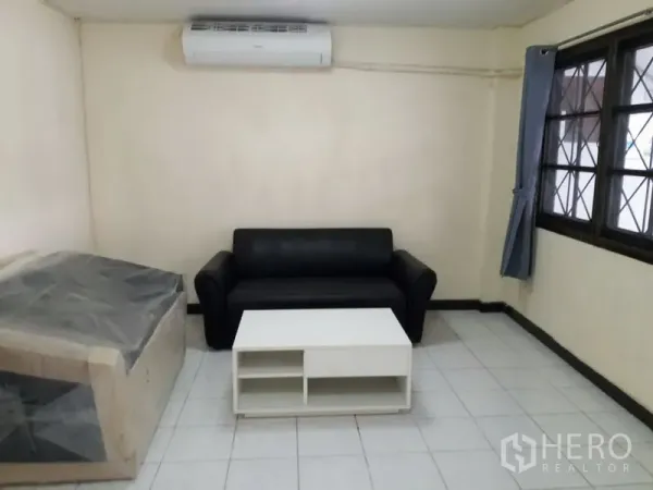 Townhouse for rent in Watthana, Bangkok - Simple living room with black sofa, white coffee table, tiled floor and wall-mounted air conditioner.