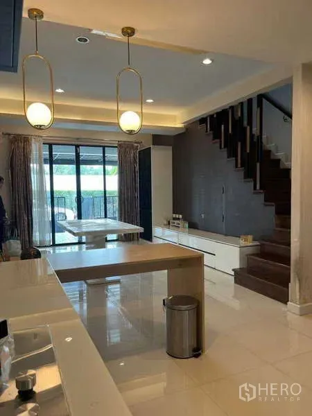 Townhouse for rent in Khlong Toei, Bangkok - Open-plan kitchen and dining area with pendant lights, island, and wooden staircase leading to patio doors.